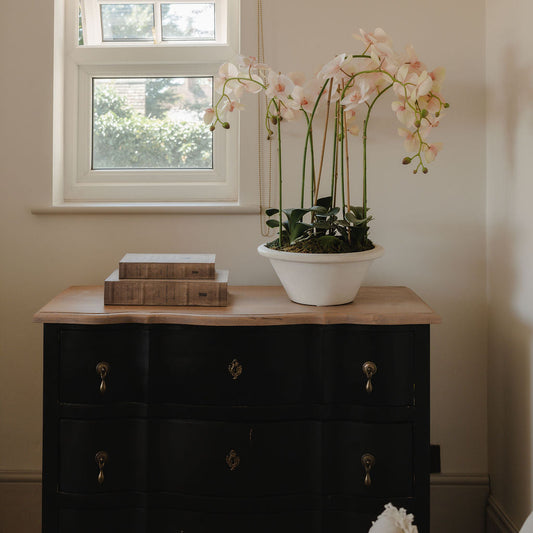 Black dresser with a white vase of flowers and books against a window in a room.