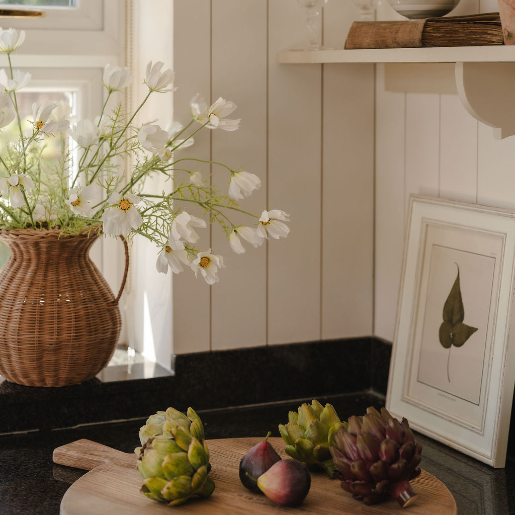 Wooden cutting board with fruits and a vase of flowers on a kitchen counter.