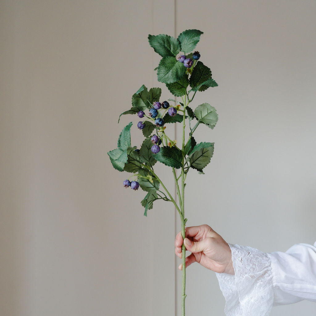 Hand holding a branch of artificial greenery with small purple flowers against a plain background