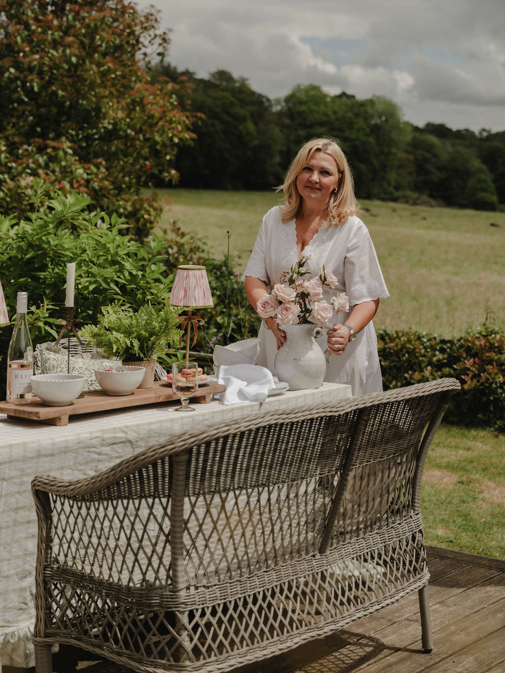 Woman standing outdoors with a table set for a meal, holding flowers.
