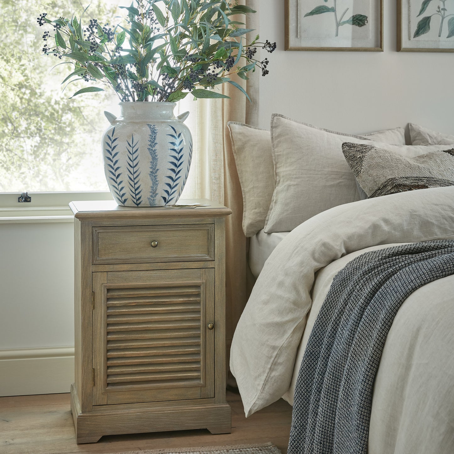 Wooden nightstand with decorative vase and plant next to a bed in a bedroom.