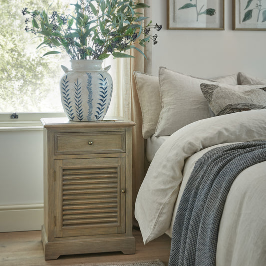 Wooden nightstand with decorative vase and plant next to a bed in a bedroom.