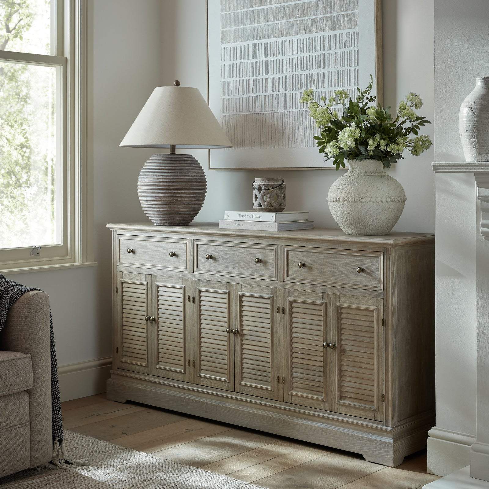 Light wooden sideboard with decorative items in a room with a window and fireplace.