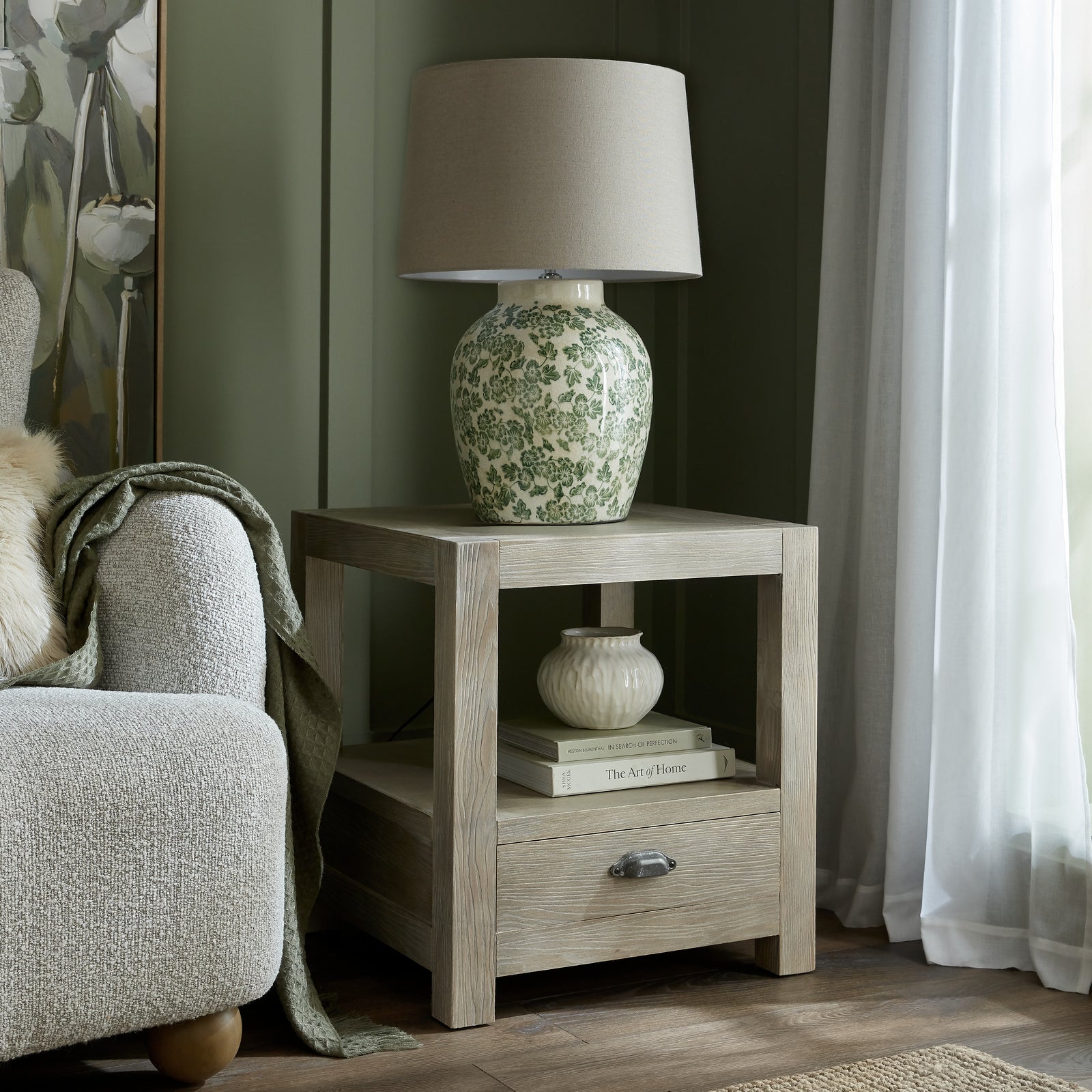 Side table with a floral-patterned lamp and decor items next to a chair in a room with curtains.