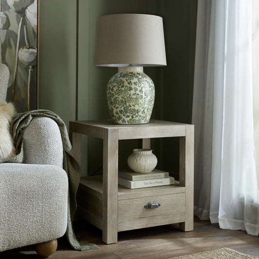 Side table with a floral-patterned lamp and decor items next to a chair in a room with curtains.