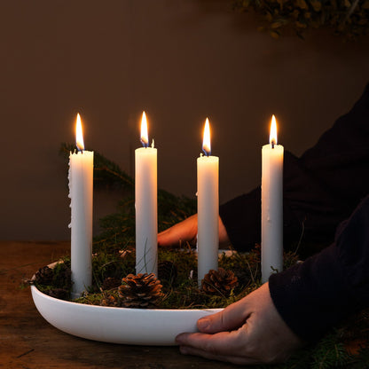 Four lit candles in a white dish with greenery on a wooden surface.