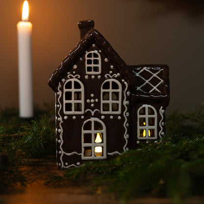 A ceramic house with a gingerbread design, decorated in black and white, sitting on a bed of greenery with a lit candle beside it, creating a Christmas decoration theme.
