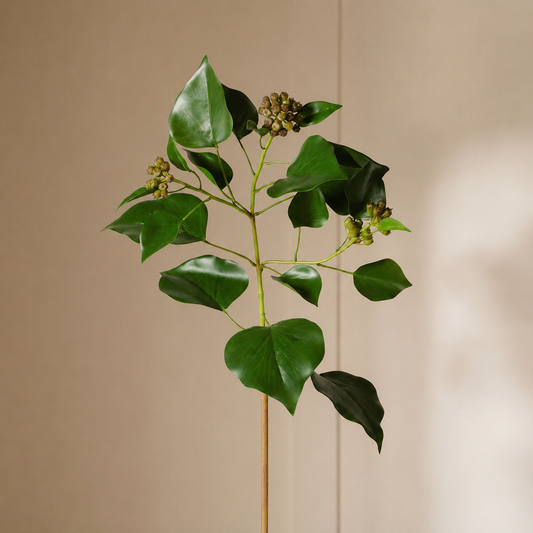 A single artificial green leafy branch with small clustered buds on a wooden stick, photographed against a warm beige wall with soft sunlight and shadows.