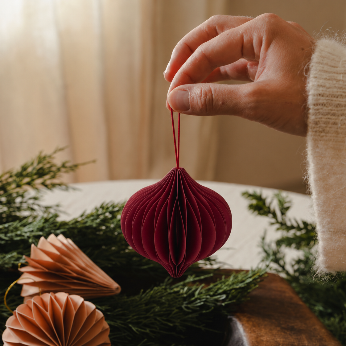 A hand wearing a cream knit sweater holds a deep burgundy paper honeycomb Christmas ornament against a warm, softly blurred background with green pine branches and neutral decor.