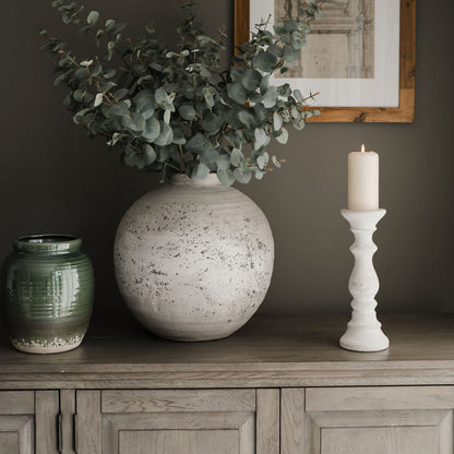 A faux variegated cineraria eucalyptus stem in sage color placed in a white vase, alongside a picture frame and a candle on a wooden cabinet.