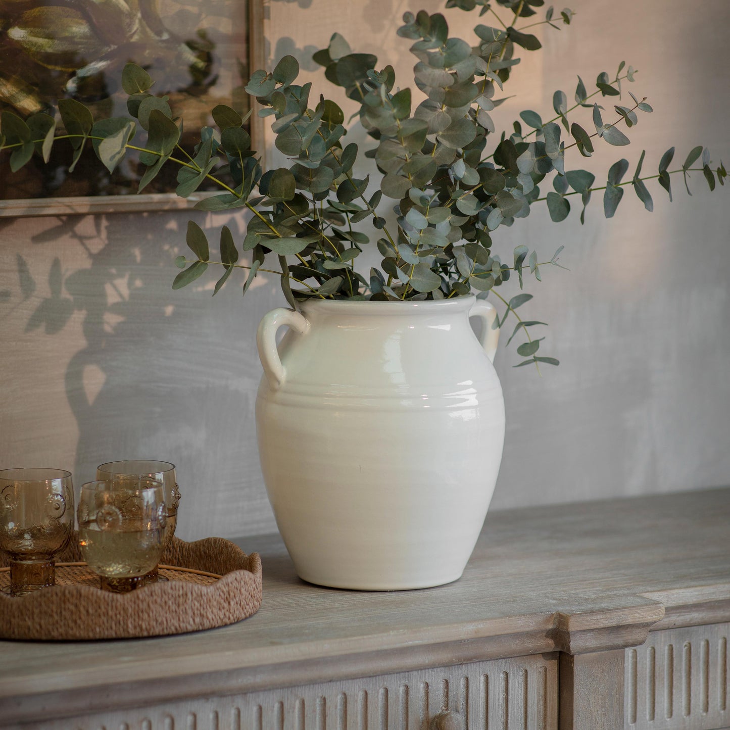 Fresh eucalyptus stems arranged in a large white ceramic vase on a rustic wooden sideboard, styled with warm neutral decor and soft ambient lighting