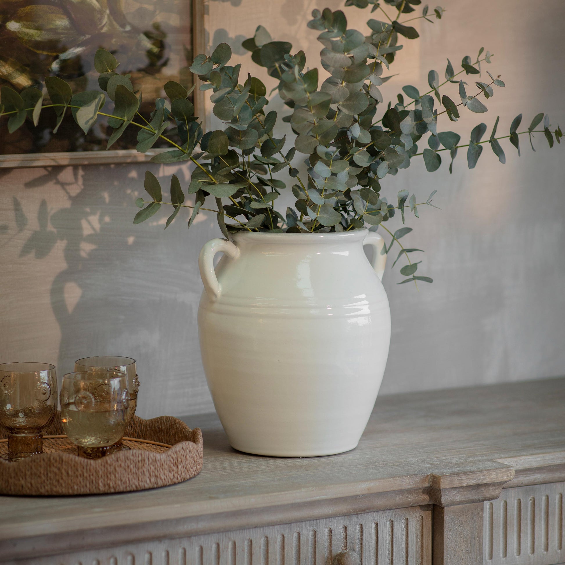 Fresh eucalyptus stems arranged in a large white ceramic vase on a rustic wooden sideboard, styled with warm neutral decor and soft ambient lighting