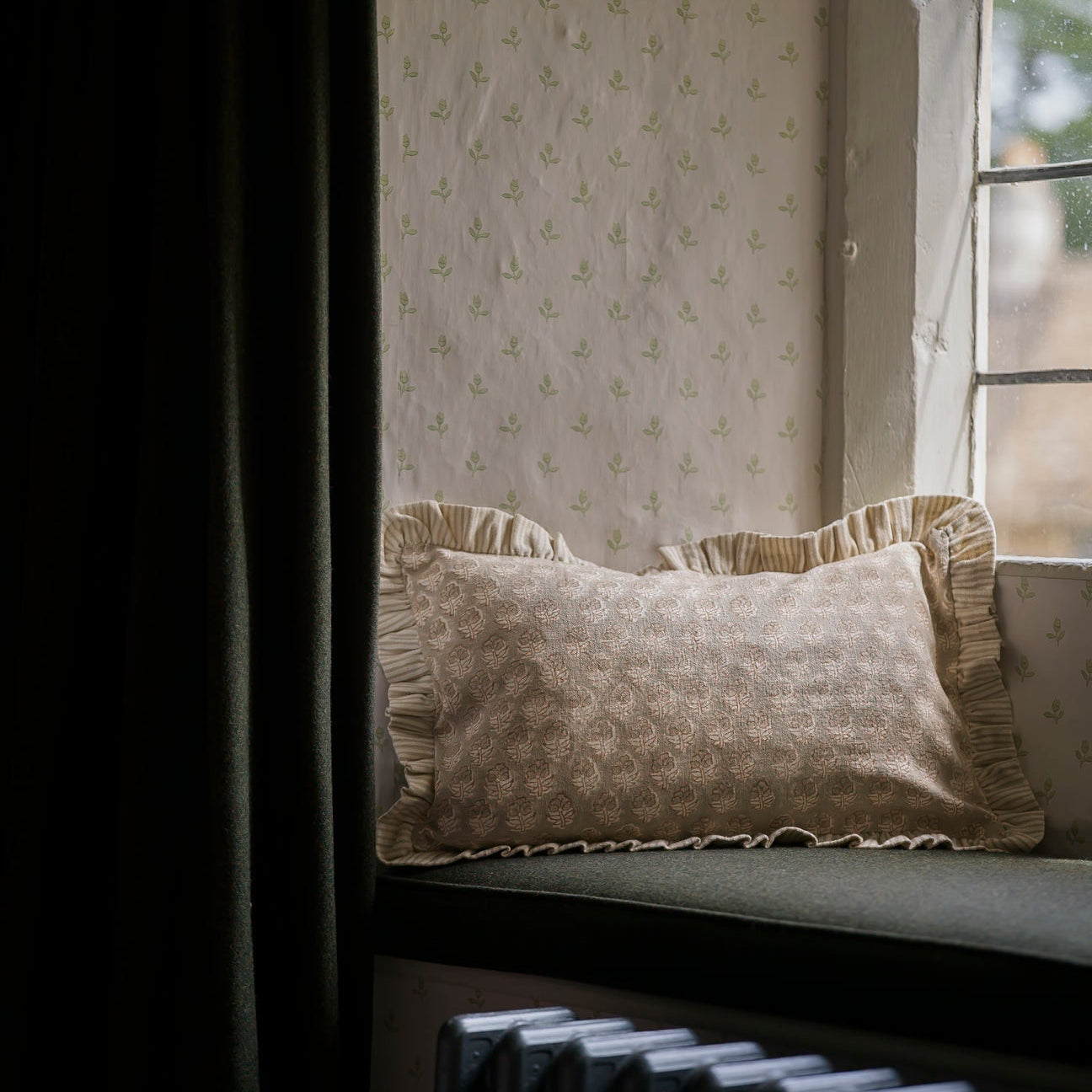 Decorative pillow on a window seat with patterned wallpaper and curtain in the background