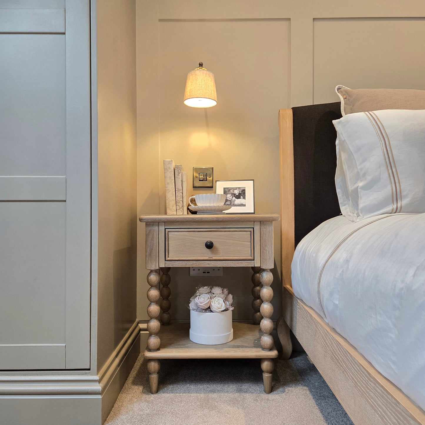 Rustic wooden bedside table with turned ball-leg detailing and a single drawer, styled with framed art, a ceramic tray, and flowers on the lower shelf beside a bed in a warm neutral bedroom