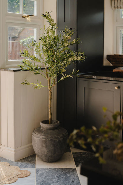 Artificial olive tree with realistic green foliage in a dark rustic planter, styled beside black kitchen cabinets and bright windows in a modern farmhouse kitchen