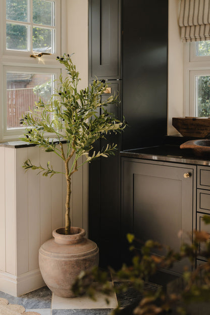 Tall artificial olive tree in a rustic clay pot styled in the corner of a modern dark kitchen with black cabinetry and natural light from surrounding windows
