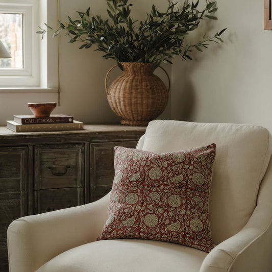 White armchair with a patterned pillow, wicker vase with greenery, and wooden cabinet in a room.