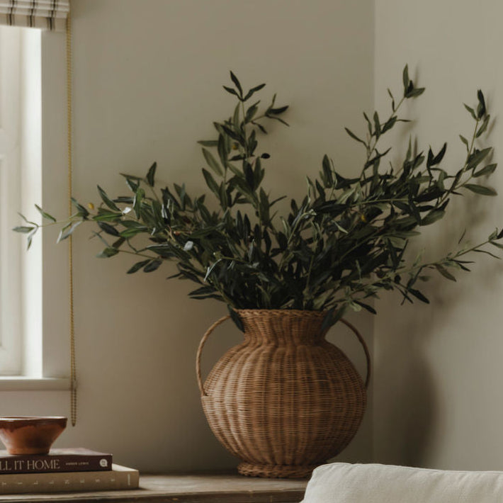 White armchair with a patterned pillow, wicker vase with greenery, and wooden cabinet in a room.