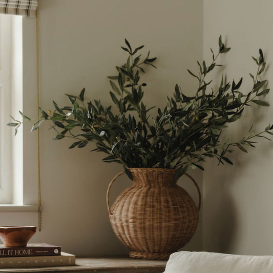 White armchair with a patterned pillow, wicker vase with greenery, and wooden cabinet in a room.