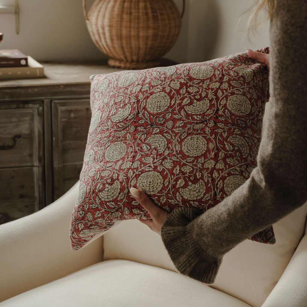 Person holding a patterned pillow in a cozy room with a vase and books in the background.