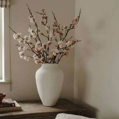 White vase with cherry blossom branches on a wooden surface against a beige wall.