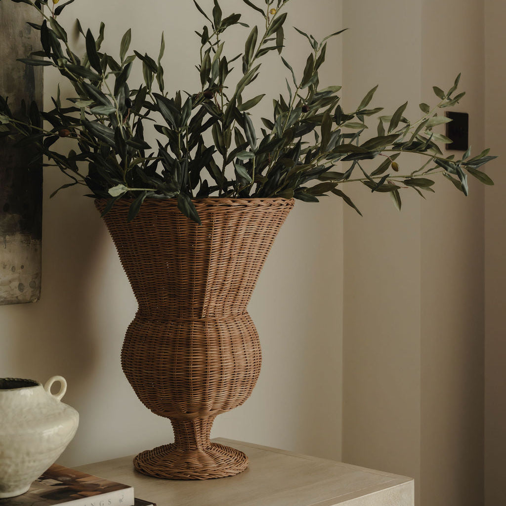 Wicker vase with green foliage on a wooden surface against a neutral wall.