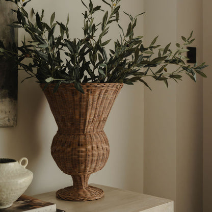 Wicker vase with green foliage on a wooden surface against a neutral wall.