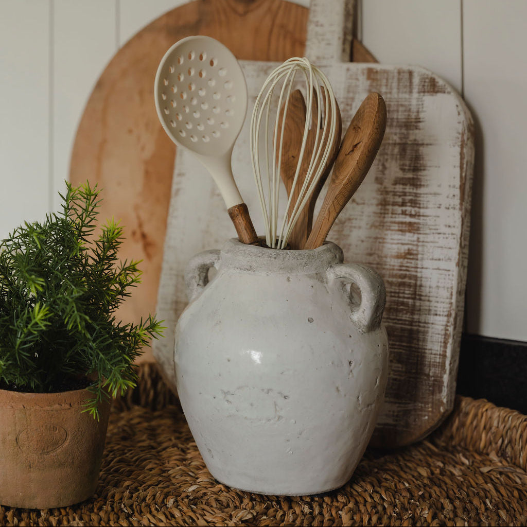 Decorative arrangement with wooden cutting boards, kitchen utensils in a white vase, and a small plant on a textured surface.