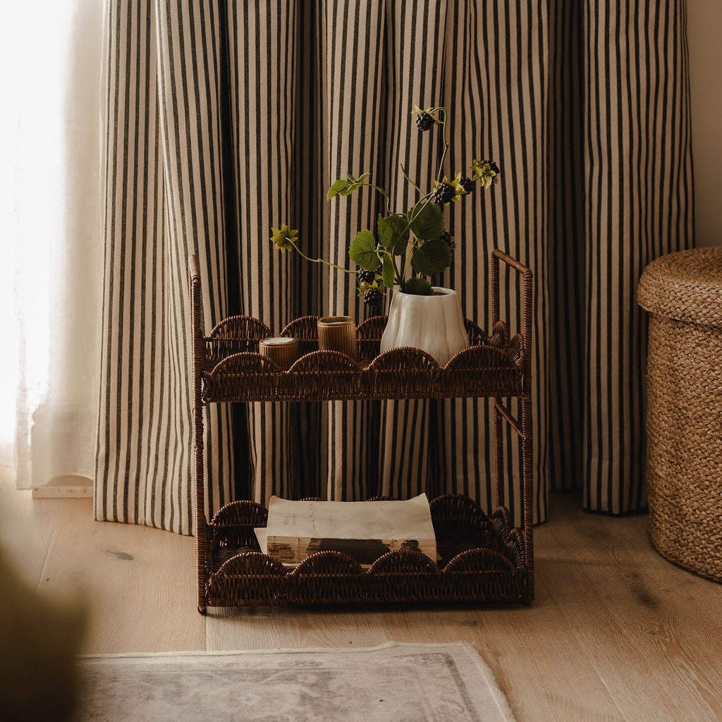 A rustic wicker two-tier cart styled in a cozy room with striped curtains, holding a white vase with green stems and small candles.