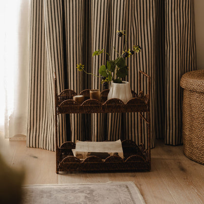 A rustic wicker two-tier cart styled in a cozy room with striped curtains, holding a white vase with green stems and small candles.