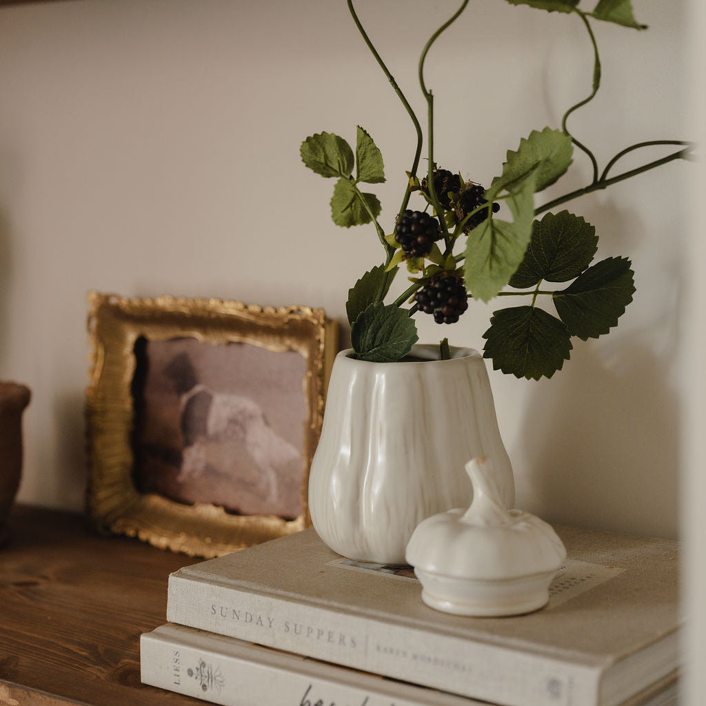 A white ribbed ceramic vase holding green leaves and dark berries, styled on a wooden shelf next to a gold picture frame and stacked books.