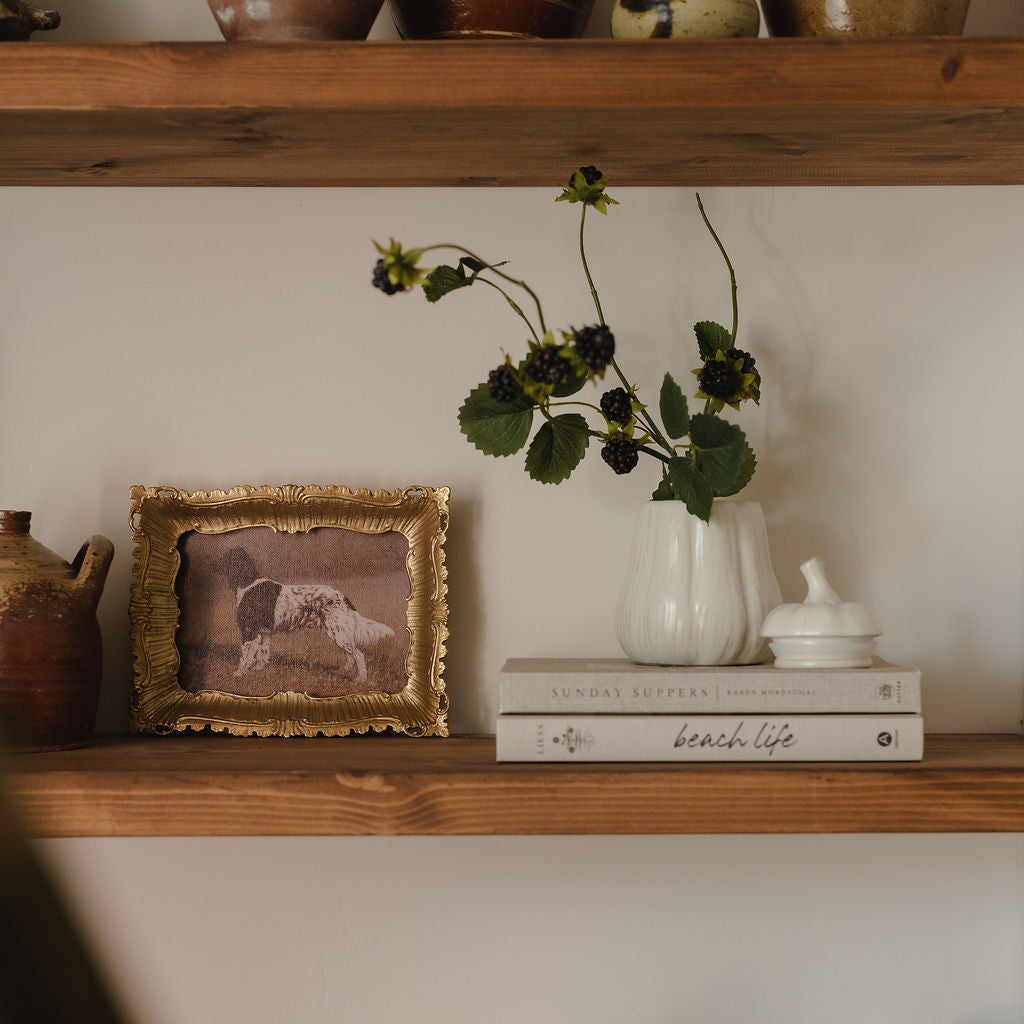 A styled interior shelf scene with wooden shelves against a white wall, holding a white vase with dried flowers, a gold-framed picture, stacked books, a ceramic jug, and books.