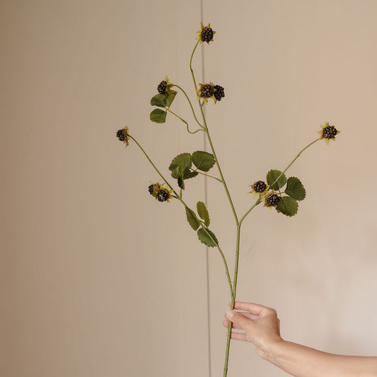 A person's hand holding a long artificial flower stem with green leaves and dark berries against a plain beige wall.