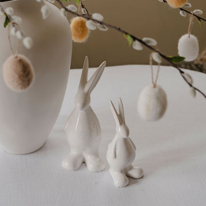 Two ceramic rabbit figures on a white surface with hanging decorative eggs and a branch in the background.