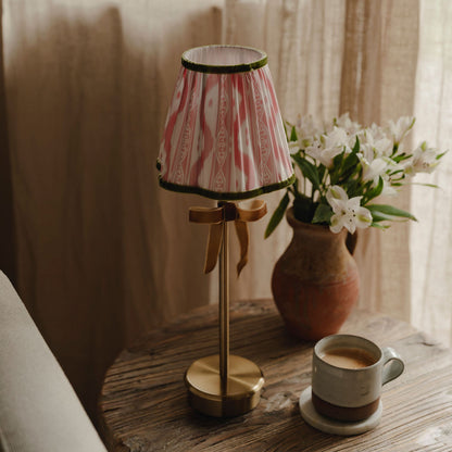 Small lamp with a striped shade on a wooden surface next to a cup of coffee and a vase of flowers.