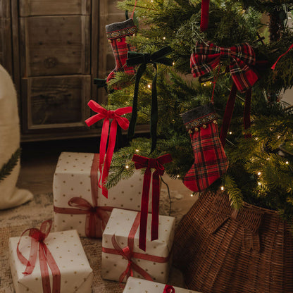 A cozy Christmas scene with a decorated tree and wrapped presents. The tree has red plaid stockings, red ribbon bows, and warm white string lights, with several gift boxes wrapped in cream paper with gold dots and red ribbons underneath.