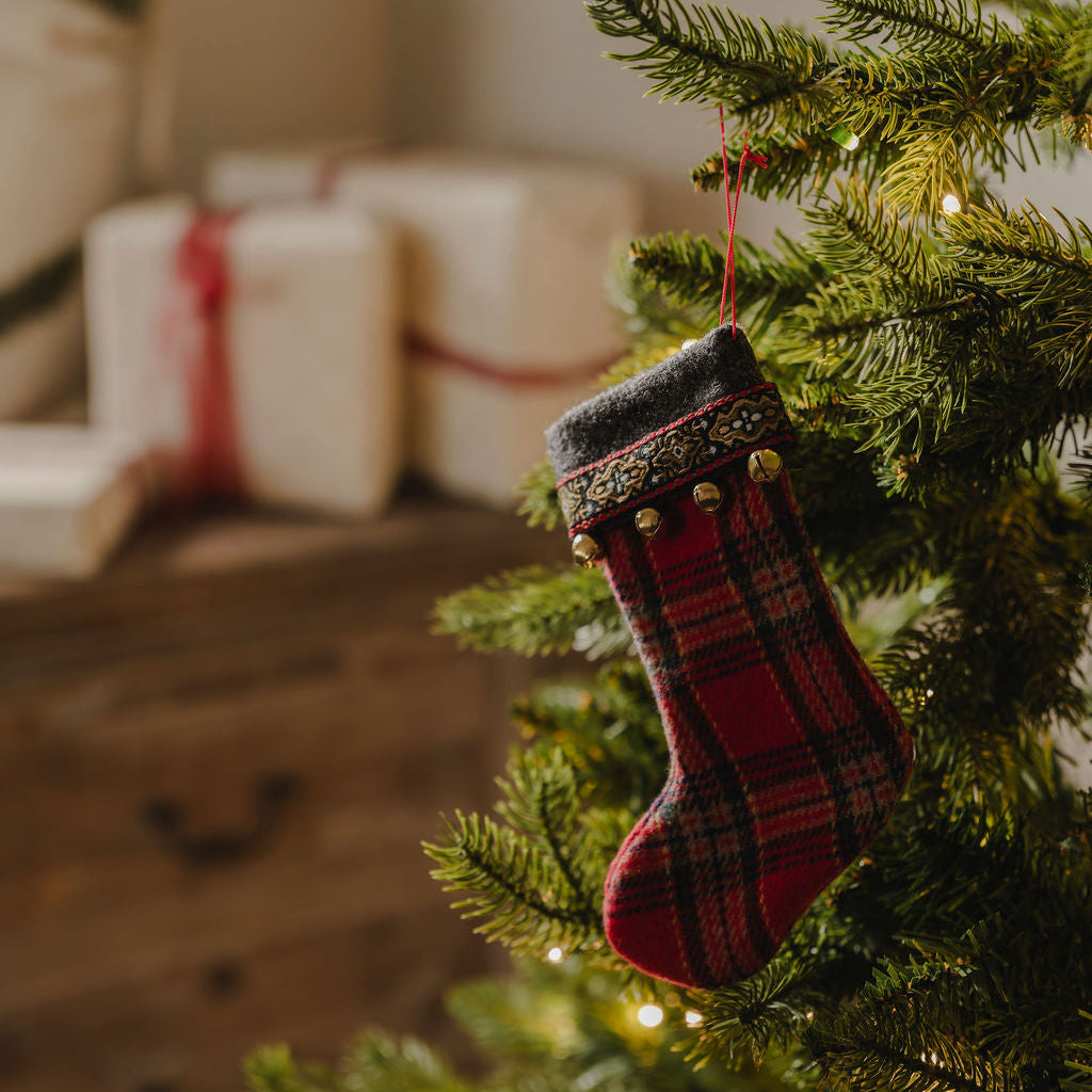 A red and black plaid Christmas stocking ornament hangs from the green branches of a decorated Christmas tree, with warm string lights and blurred gift boxes in the background.