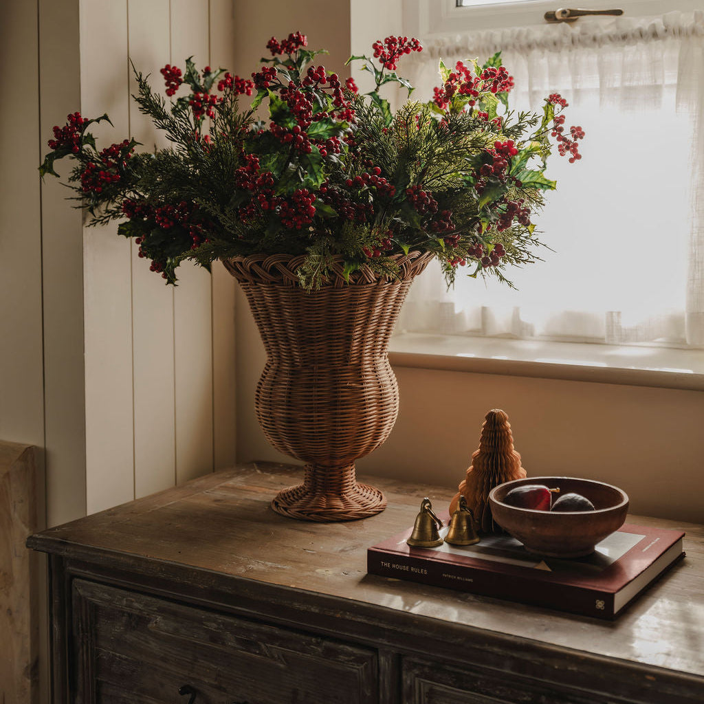 A large wicker urn-shaped vase filled with red berries and green foliage sits on a rustic wooden console table in a sunlit room near a window.