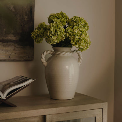 White vase with green flowers on a wooden surface against a neutral wall.