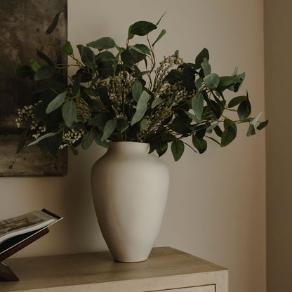 Minimalist white ceramic vase with lush green eucalyptus stems and delicate white filler flowers on a wooden table against a warm neutral wall