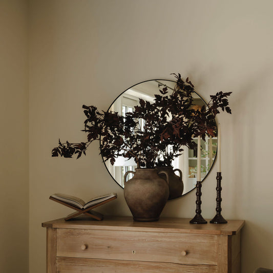 A rustic light brown wooden chest of drawers in a styled room, topped with a large round mirror, a brown ceramic vase filled with dark foliage, an open book on a stand, and two black candlesticks.