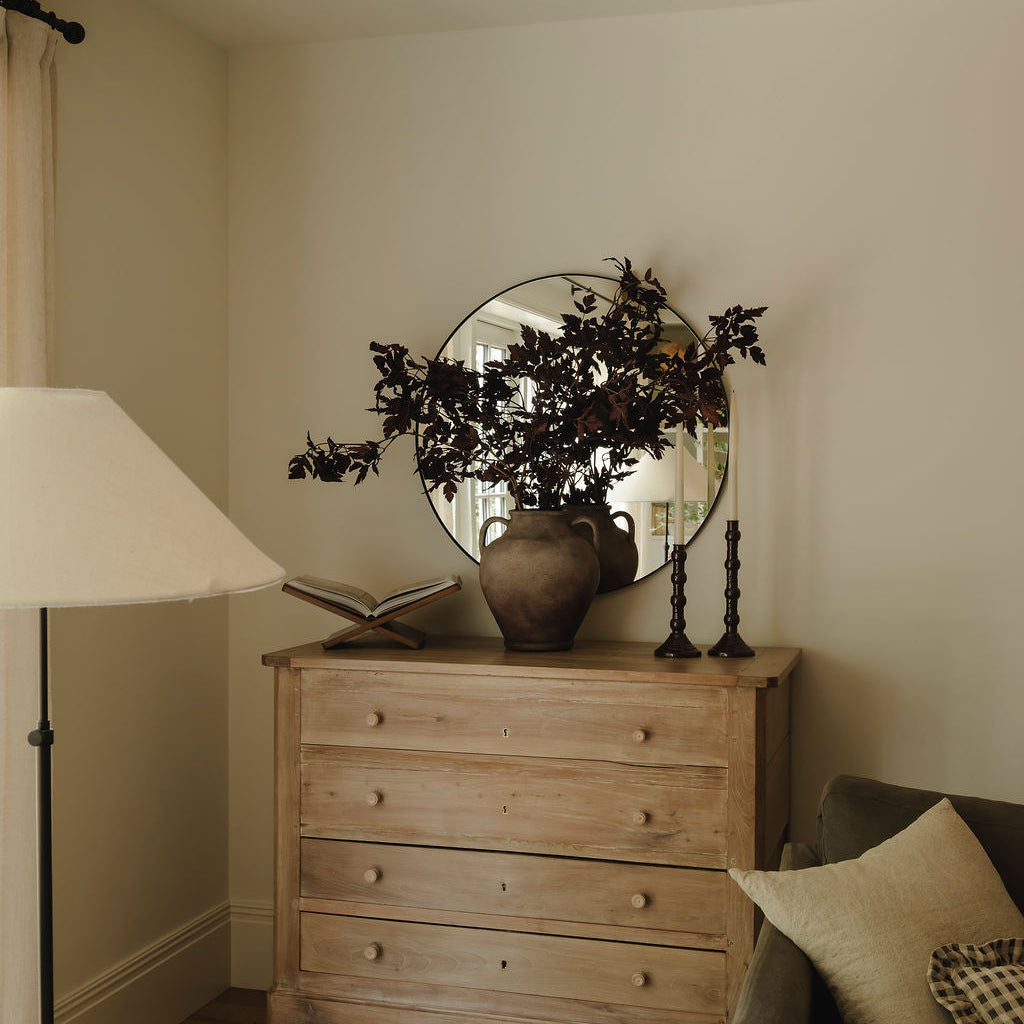 A rustic light wood chest of drawers in a cozy room setting, styled with a round mirror, a ceramic vase of dark leaves, an open book, and candlesticks.