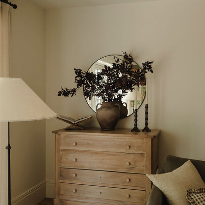A rustic light wood chest of drawers in a cozy room setting, styled with a round mirror, a ceramic vase of dark leaves, an open book, and candlesticks.