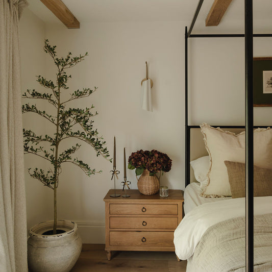 A styled bedroom corner featuring a light wood three-drawer nightstand with a vase of dark burgundy flowers, a potted tree, and a black metal canopy bed with white bedding.