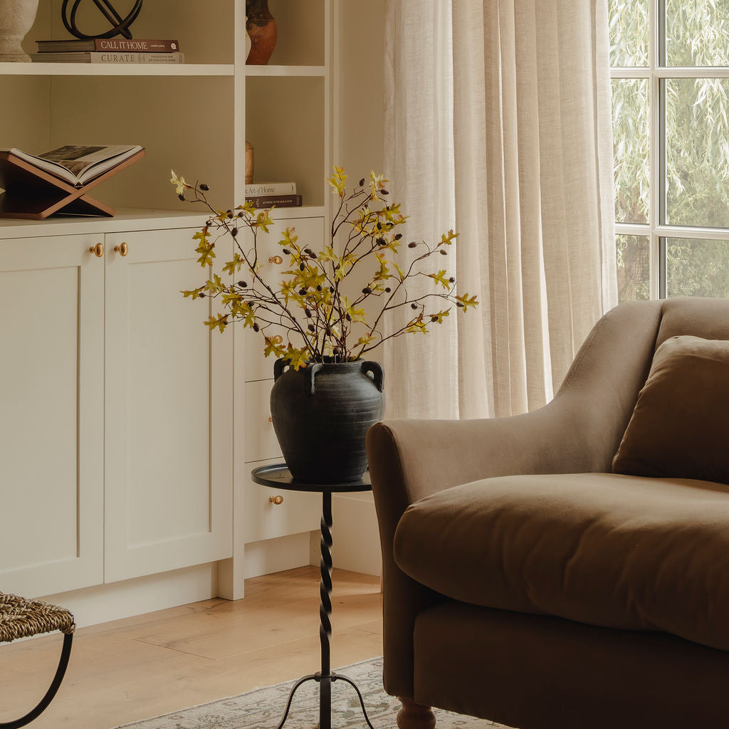 A cozy living room corner featuring a brown upholstered armchair, a black side table with a dark ceramic vase of yellow blossoms, cream curtains, and light wood flooring.