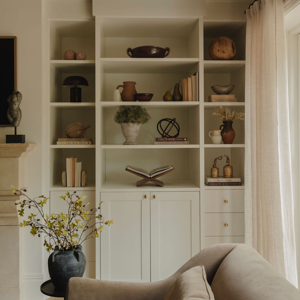 A styled interior scene showing a large built-in white shelving unit with decorative books, vases, and plants, set in a warm living room with a beige armchair in the foreground and a window with sheer curtains on the right.