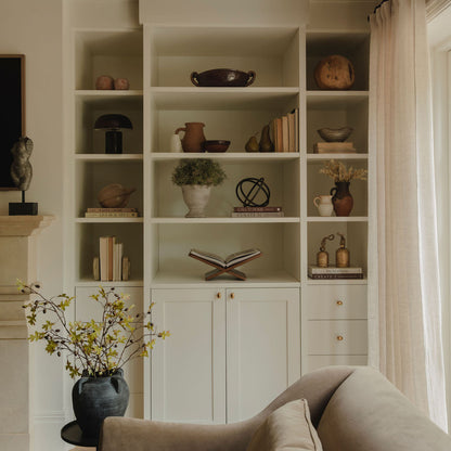 A styled interior scene showing a large built-in white shelving unit with decorative books, vases, and plants, set in a warm living room with a beige armchair in the foreground and a window with sheer curtains on the right.