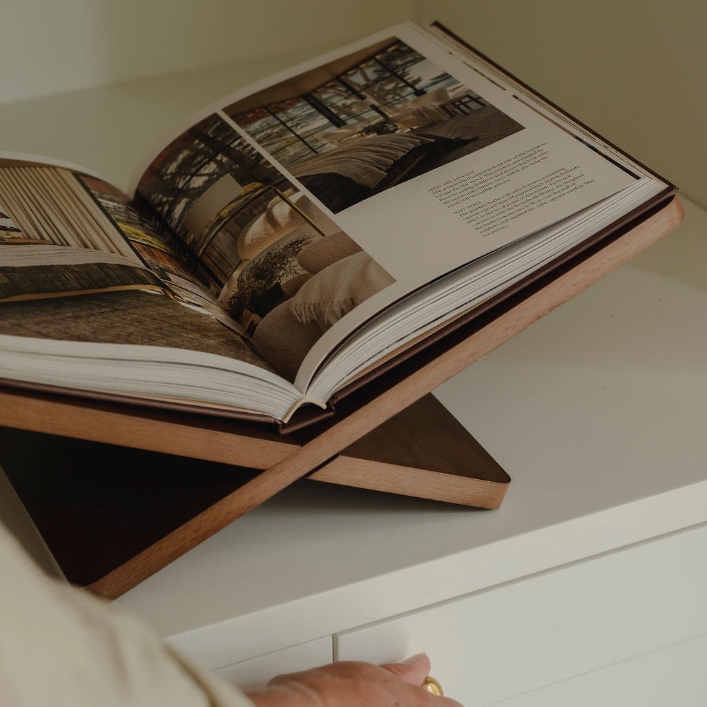 An open architectural or design book displayed on a wooden shelf riser, with a person's hand and arm visible holding the lower shelf open. The scene is photographed in a clean, modern interior with warm, neutral lighting.