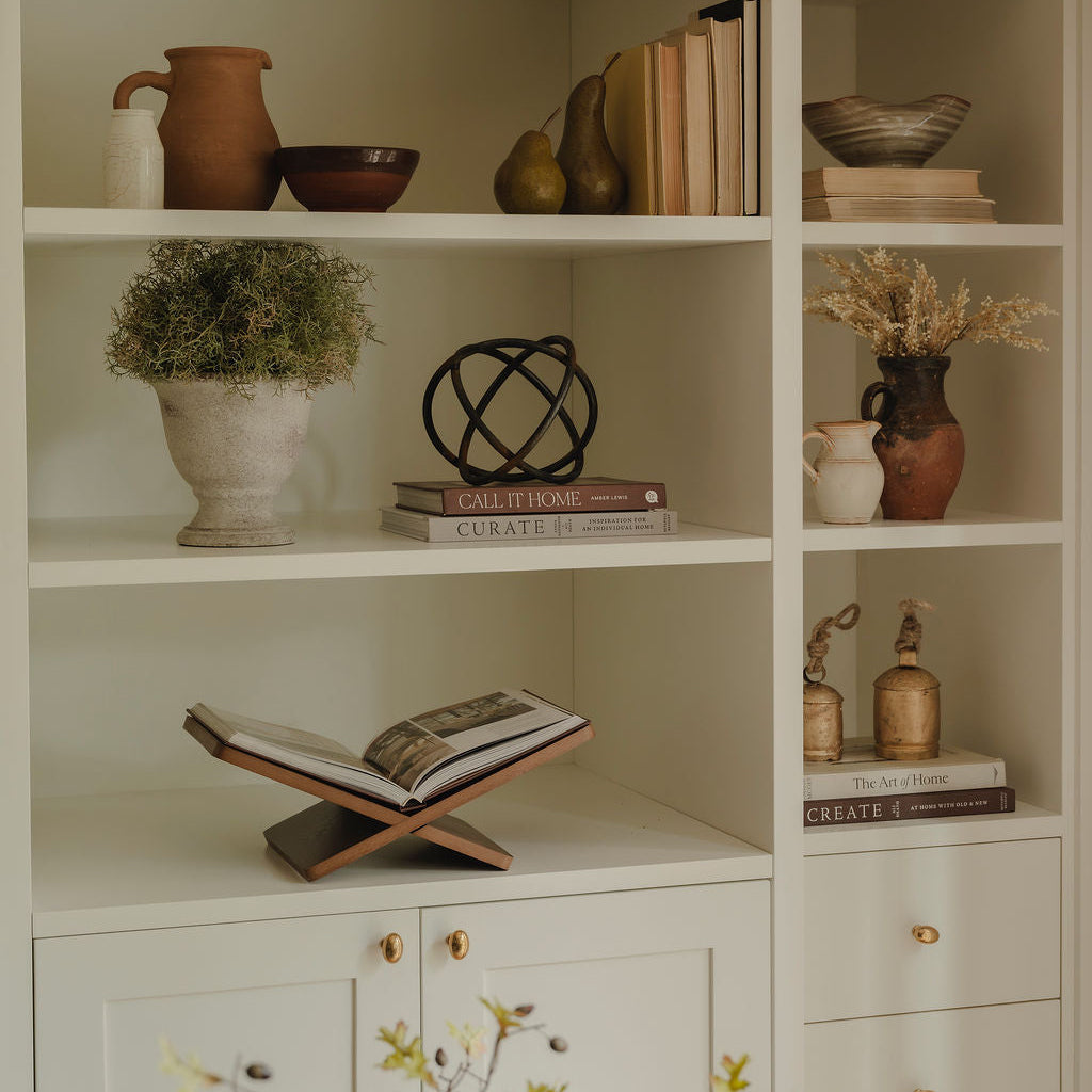A tall built-in shelving unit in a warm cream or off-white tone, styled with books, ceramic vases, bowls, a plant in a pot, dried flowers, and an open book on a wooden stand.