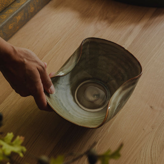 A hand holds a handmade ceramic bowl with a wavy rim and a textured, earthy green-brown glaze, set on a light wooden table with books and a tray in the background.